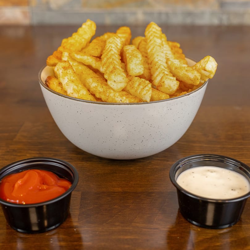 A white bowl of seasoned crinkle-cut fries with small black cups of ketchup and ranch dressing.