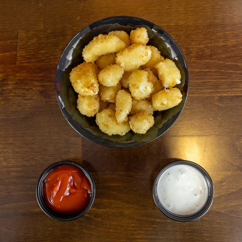 A bowl of golden-brown fried cheese curds with sides of ketchup and ranch dressing on a wooden table.