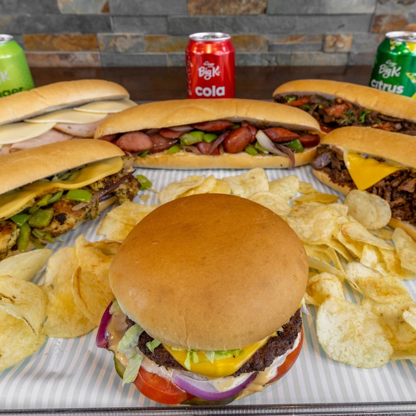 A burger, four sub sandwiches, potato chips, and three Big K soda cans on a striped tray.